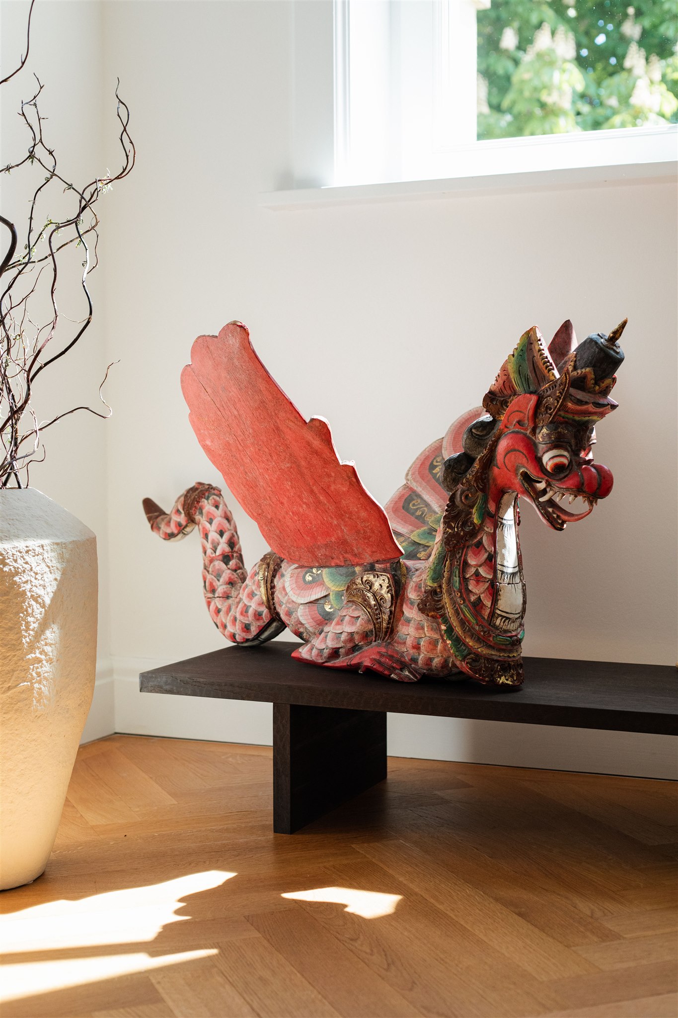 Eine kunstvoll geschnitzte, bunte Drachenfigur mit großen roten Flügeln, die auf einem schwarzen Holztisch im Raum mit Holzboden und weißen Wänden sitzt.